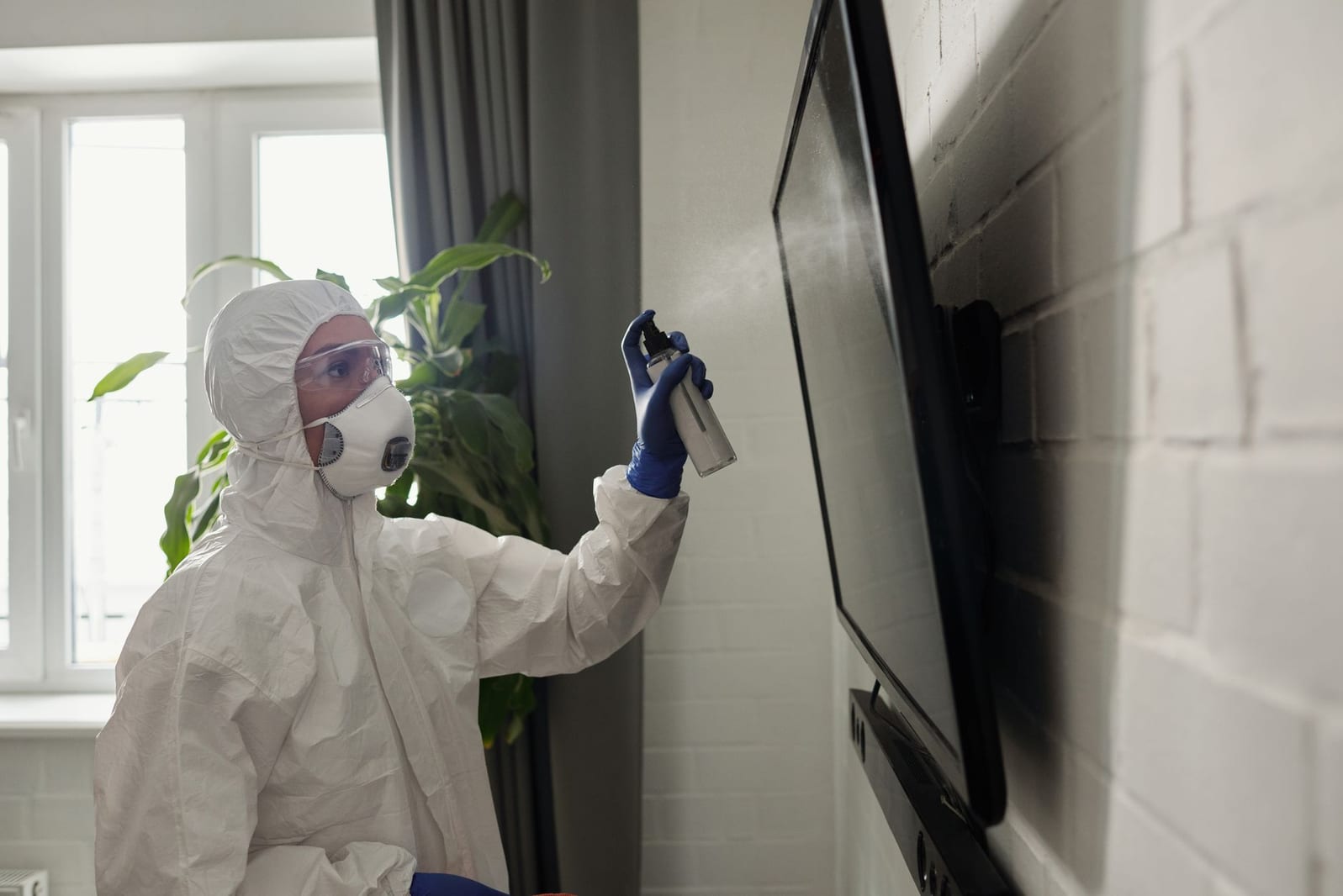 A pest-control technician in goggles and respirator applying a targeted spray along an apartment wall